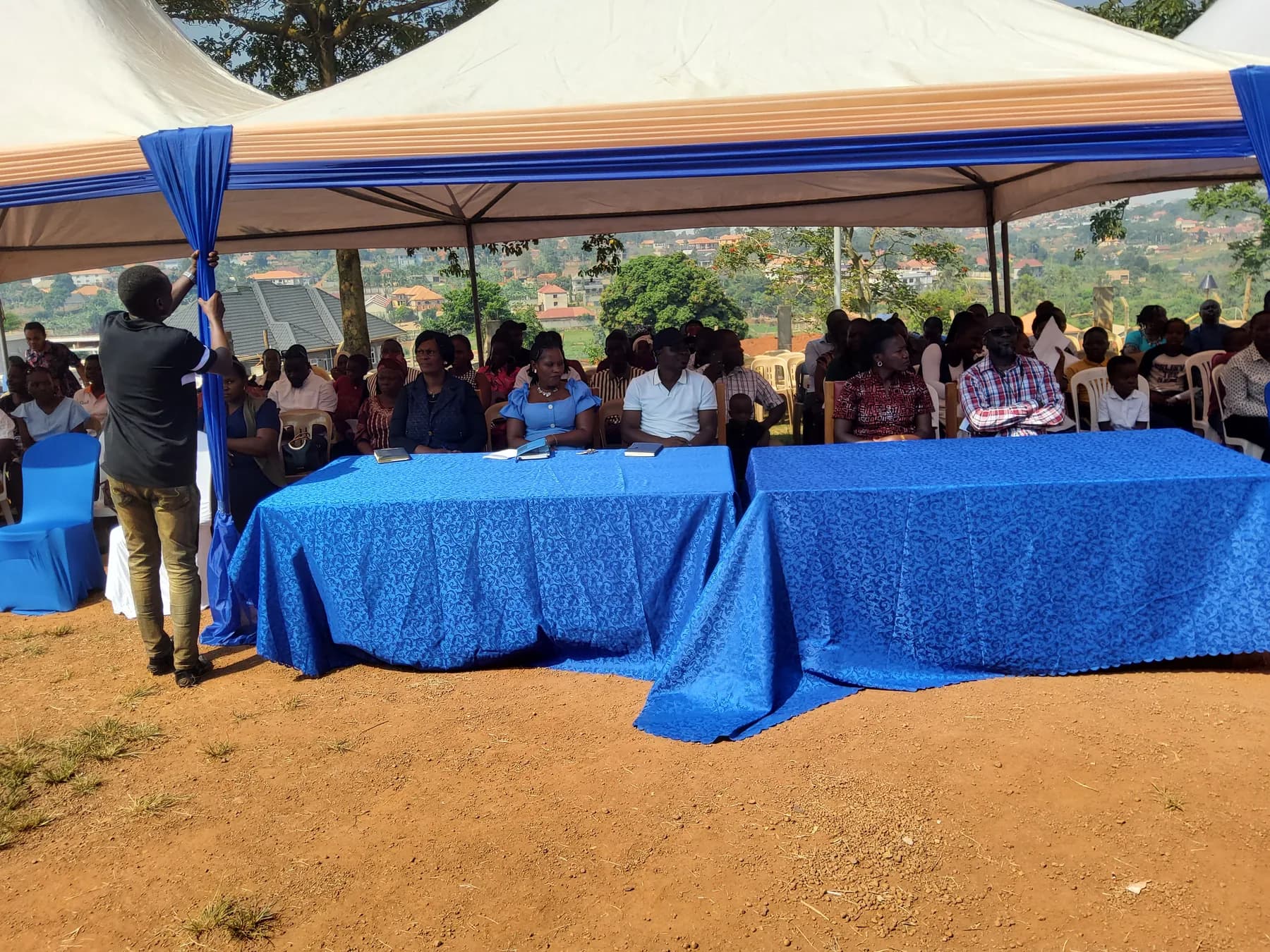 Families and invited guests seated behind blue-covered tables under a decorated tent