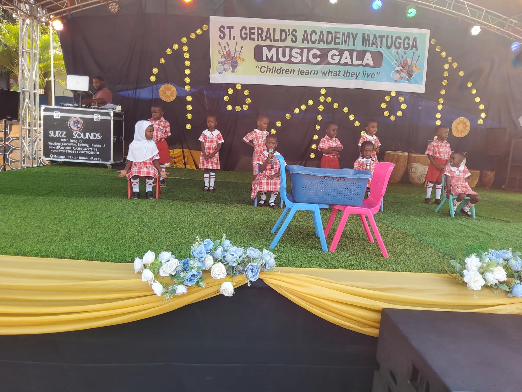 Young learners in red checked school uniform performing on stage with small chairs and a basket