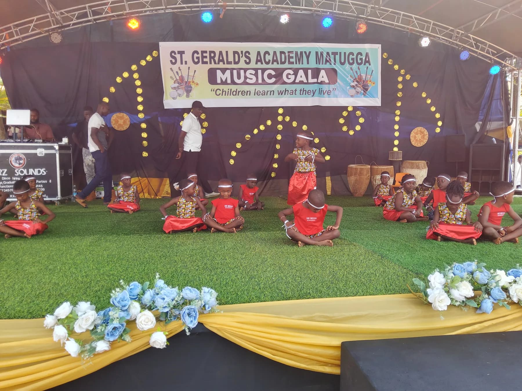 A child dancing at the centre of classmates seated in coordinated red costumes