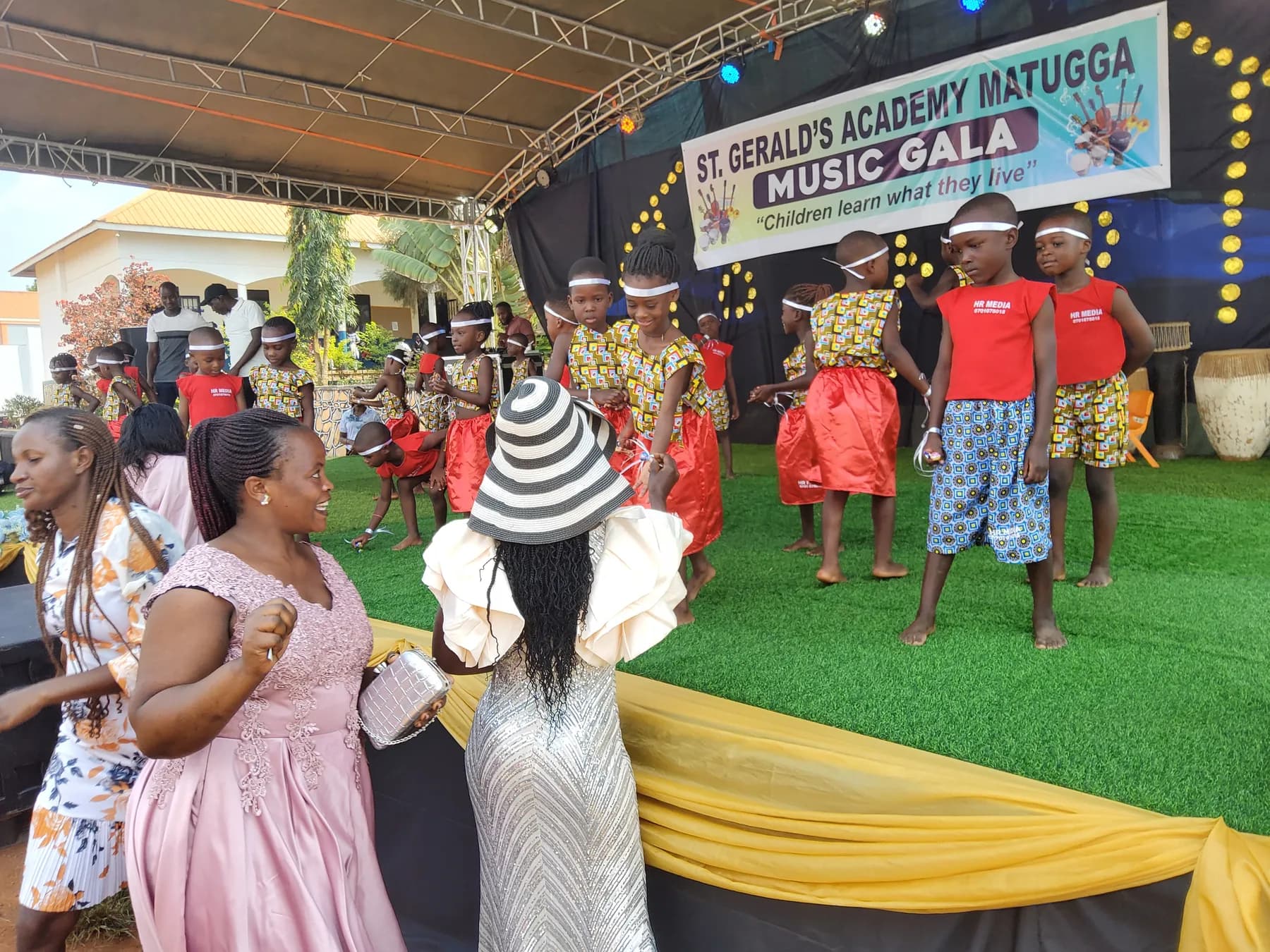 Children in red patterned performance costumes at the front of the stage as audience members greet them