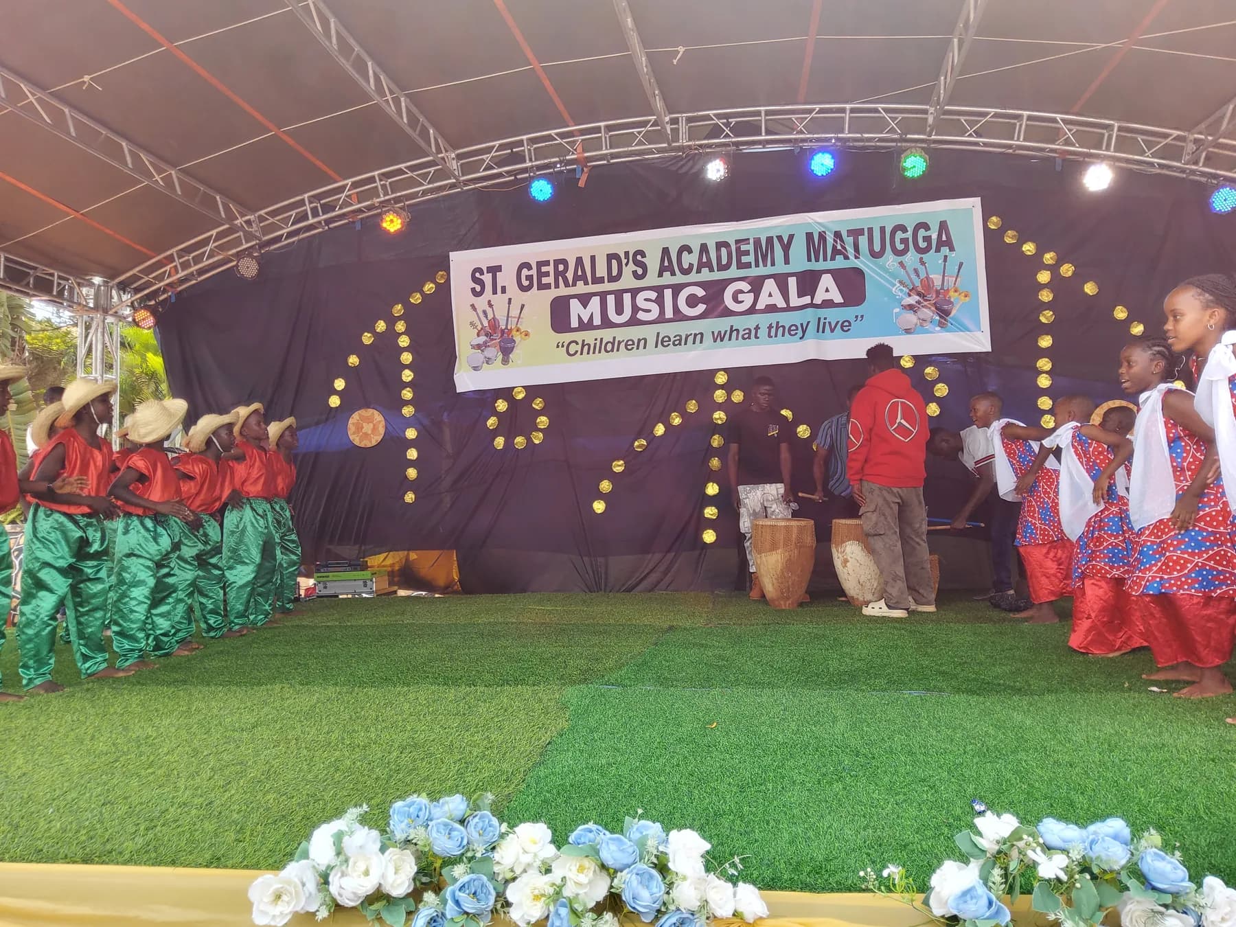 Learners in green trousers and straw hats facing dancers in red, white and blue costumes