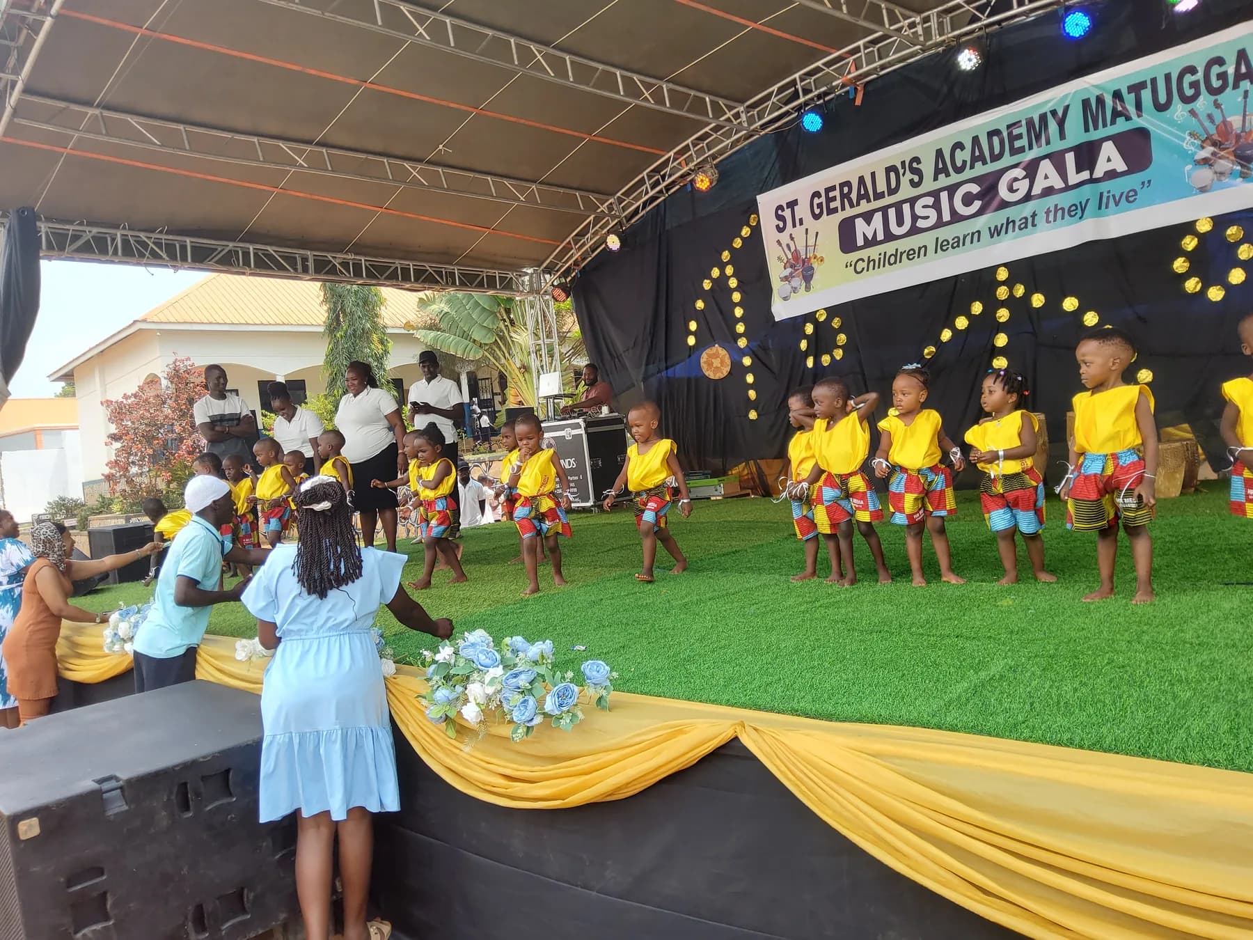 Young children in matching yellow-and-patterned outfits moving into a line across the stage