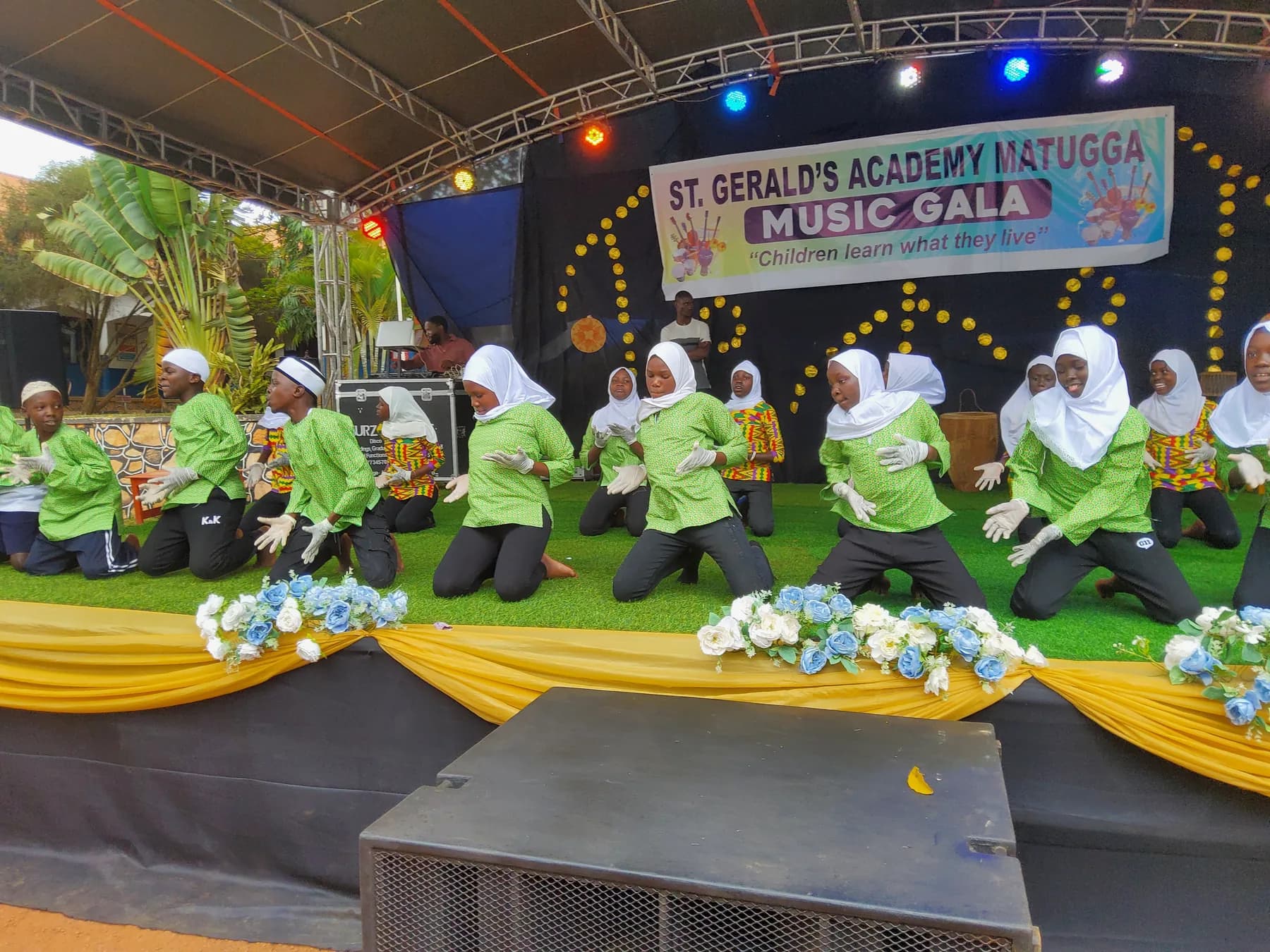 Green-and-white-costumed learners kneeling in formation and performing with expressive white-gloved hands