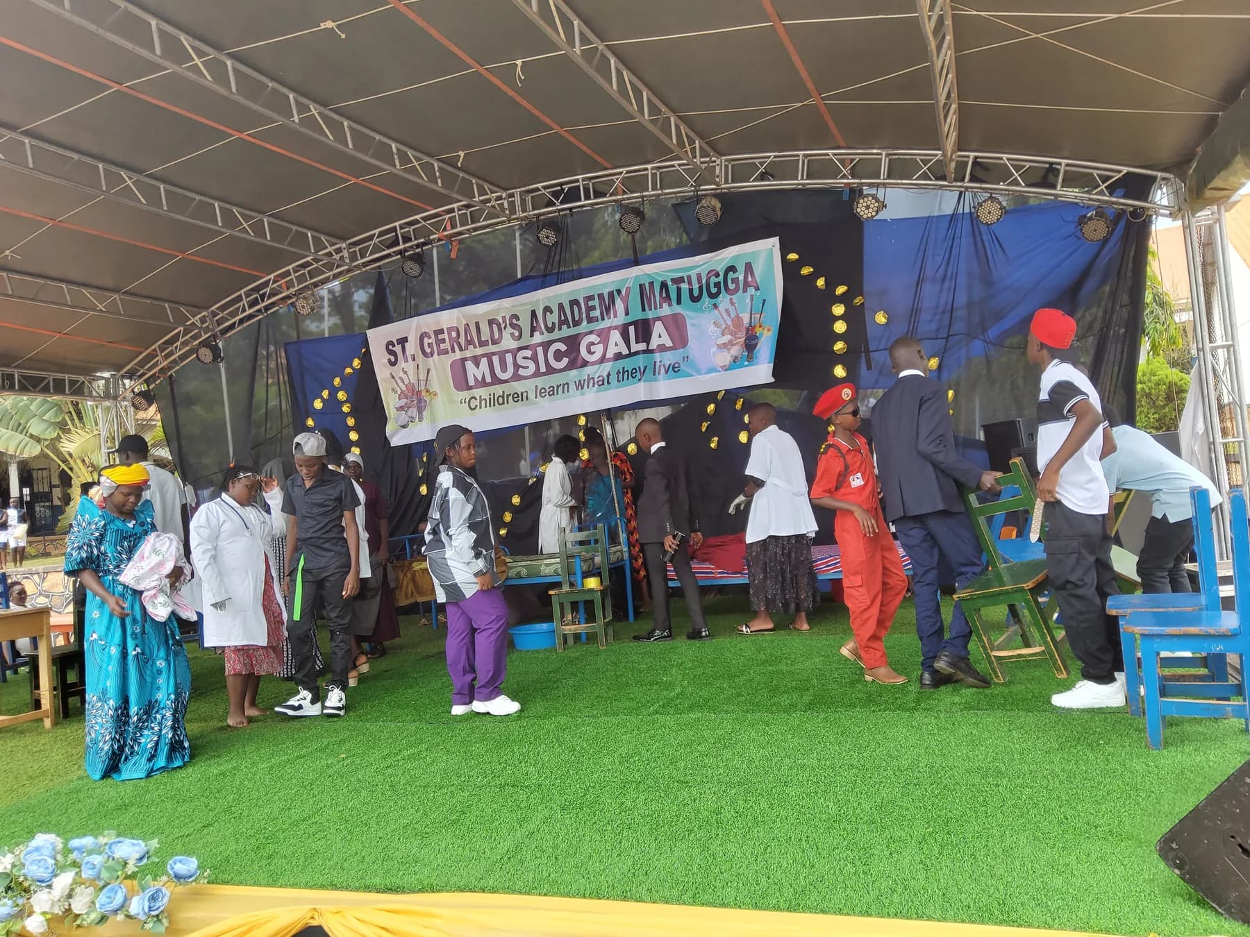 A large drama cast in varied costumes moving among beds and chairs on stage