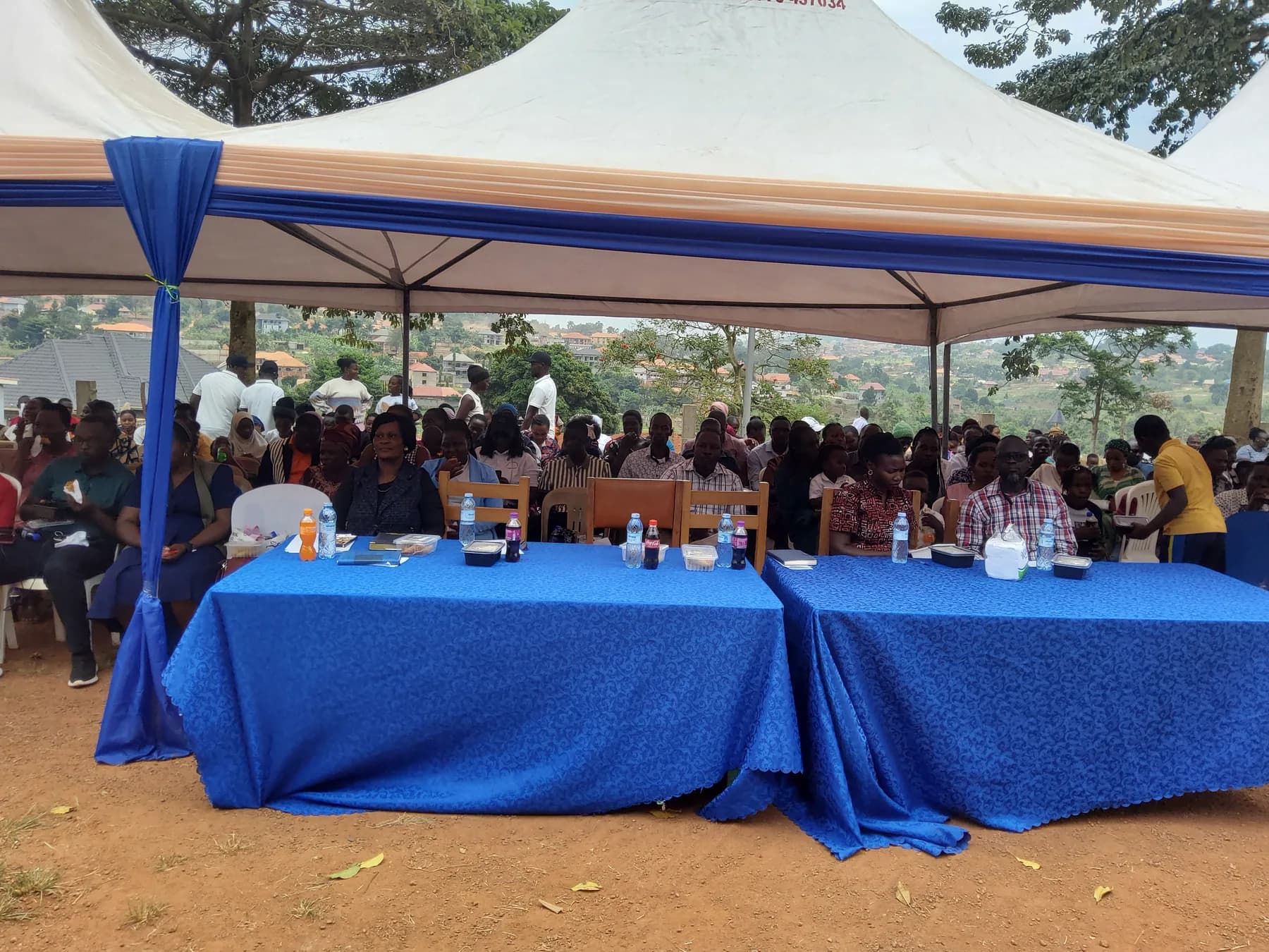 Guests seated behind blue-covered front tables with the larger audience beneath a tent behind them