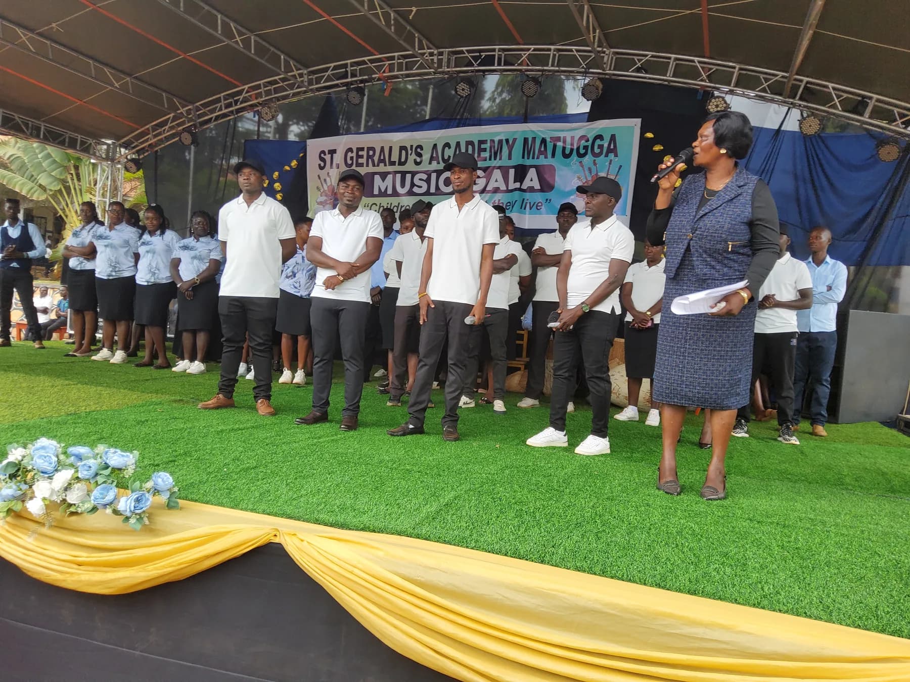 A speaker addressing the audience beside male staff members in white shirts and black caps