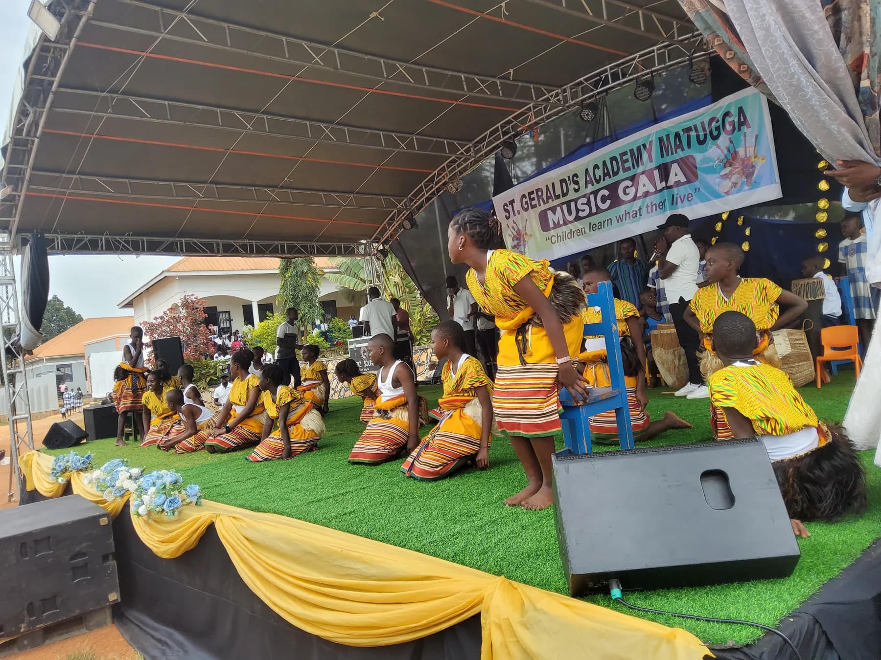 Traditional dancers in yellow and striped costumes holding a low formation around a blue chair